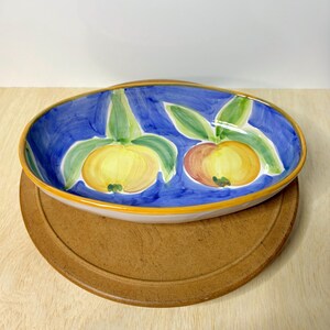 May include: An oval ceramic serving dish featuring a hand-painted design of two yellow fruits with green leaves against a vibrant blue background. The dish has a distinct yellow rim and a smooth, glossy finish, showcasing a classic artisan style.