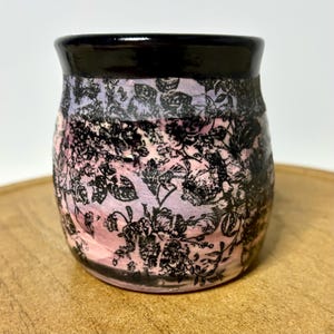 May include: A ceramic tumbler with a black rim and a floral pattern. The body of the tumbler transitions from pink to lavender, with black floral designs. The tumbler is sitting on a wooden surface.