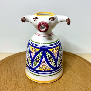 May include: A white ceramic bull-shaped vase with a red mouth and brown horns. The vase has a blue and yellow patterned design around the base and a yellow rim. The vase is sitting on a wooden surface.