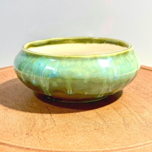 May include: A round ceramic bowl featuring a glossy glaze in shades of light green, mint, and soft blue. The exterior surface displays a distinctive drip pattern where the glaze runs downward, while the interior is a smooth, solid off-white color.