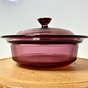 May include: A vintage, ruby-colored glass casserole dish with a lid. The dish has a ribbed design on the sides and a rounded lid with a small handle. The dish is sitting on a wooden surface.