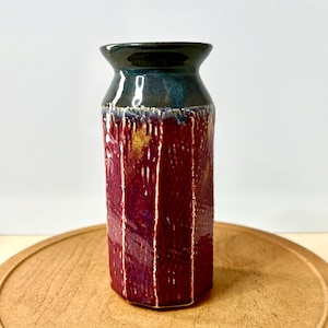 May include: A ceramic vase with a dark teal rim and a deep burgundy body. The vase has a textured surface with vertical lines and a hexagonal base. The vase sits on a wooden surface.