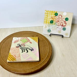 May include: Two decorative square ceramic tiles. One tile features a fox illustration with a patchwork design. The other tile has a floral design with green and red accents. Both tiles are on a wooden surface.