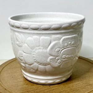 May include: A white ceramic flower pot with embossed floral and leaf designs. The pot has a rounded shape with a decorative beaded rim. The pot is sitting on a wooden surface.