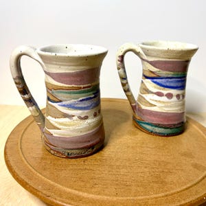 May include: Two ceramic mugs with speckled off-white surfaces featuring horizontal bands of muted purple, blue, and green glaze. Each mug has a curved handle and a flared rim. The base of the left mug displays the embossed text KICKAPOO.