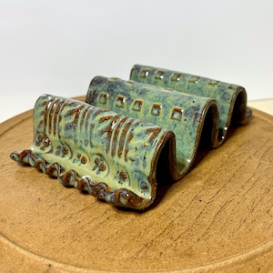May include: A ceramic soap dish with a wavy design. The dish is glazed in shades of green and brown, with a textured surface. The dish is designed to hold multiple bars of soap.