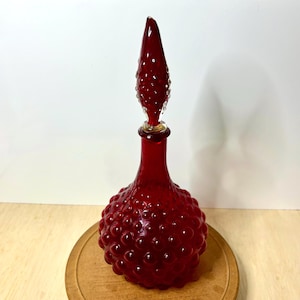 May include: A ruby red glass decanter with a textured, bubble-like design on the body. The stopper is elongated and pointed, with a textured surface and clear glass accents. The decanter sits on a wooden surface.