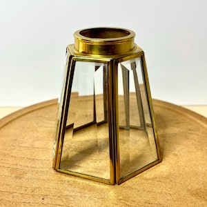 May include: A clear glass and brass-toned geometric candle holder. The holder has a hexagonal base and a cylindrical top. The brass-toned metal frames the clear glass panels, creating a vintage or antique aesthetic. The candle holder is on a wooden surface.