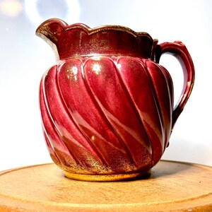 May include: A ceramic pitcher featuring a deep red glaze with a metallic gold finish on the rim, handle, and base. The body of the pitcher has a distinct swirling, ribbed texture and a scalloped edge around the top opening.