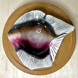 May include: A ceramic fish-shaped dish with a brown, burgundy, and white color scheme. The dish is placed on a round, brown plate. The fish has detailed fins and a glossy finish, suitable for serving appetizers or small dishes.