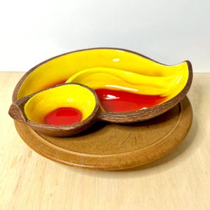 May include: A ceramic divided serving dish with a brown, wood-textured exterior and a vibrant yellow and red interior. The dish features two distinct sections, one larger and leaf-shaped and one smaller and circular, resting on a round base.