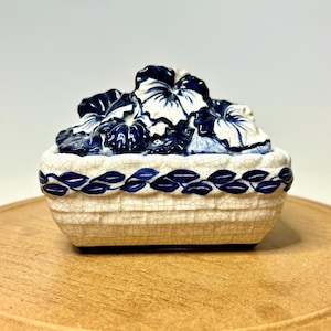 May include: A ceramic trinket box with a lid featuring a floral design. The box is rectangular with a cream-colored, crackled finish and a blue leaf pattern around the rim. The lid is adorned with blue and white flowers.