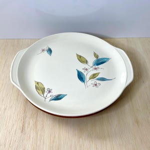May include: A round white serving platter with integrated side handles. The surface features a botanical pattern of stylized leaves in shades of blue and olive green, accented by small, delicate pink-outlined flowers scattered across the plate.