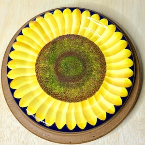 May include: A round decorative plate featuring a vibrant sunflower design. The center is a textured dark brown and green circle, surrounded by numerous bright yellow petals radiating outward against a deep navy blue background.