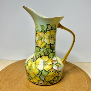 May include: A hand-painted porcelain pitcher with a curved handle and a flared spout. The pitcher is decorated with yellow rose blossoms and green leaves against a pale green background. The handle and rim are trimmed with gold.