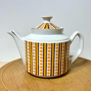 May include: White ceramic teapot with a lid, featuring a pattern of vertical orange and brown stripes with orange flower accents. The lid and spout have gold trim. The teapot is on a wooden surface.