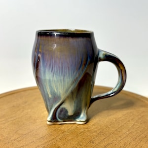 May include: A ceramic mug featuring a glossy glaze with swirling colors of deep purple, dark blue, and creamy white. The mug has a rounded body that tapers toward a square base and includes a curved handle on the right side for holding the vessel.