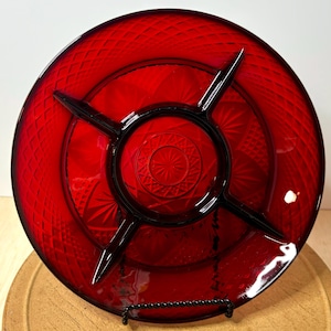 May include: A ruby red glass serving dish with a divided design. The dish features a central circular section with a starburst pattern, surrounded by three pie-shaped compartments. The outer rim has a diamond pattern.