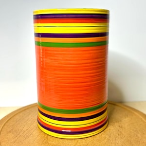 May include: A cylindrical ceramic vase featuring horizontal stripes in vibrant shades of orange, yellow, purple, and green. The surface has a glossy finish with a wide central orange band and thinner alternating colored bands throughout the body.