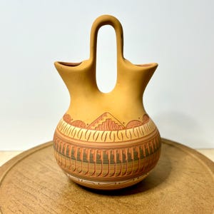 May include: A tan-colored, handcrafted pottery vase with a distinctive handle. The vase features intricate carved designs, including geometric patterns and a mountain motif. The vase is displayed on a wooden surface.