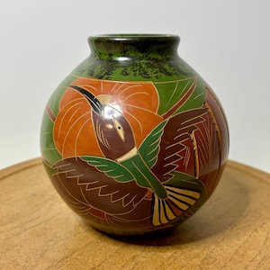 May include: A small, round ceramic vase with a detailed hummingbird design. The vase features a dark green rim, with a vibrant orange and green floral pattern. The hummingbird is depicted in shades of brown and green, with yellow accents.