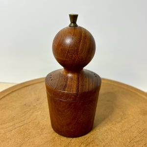 May include: A vintage wooden salt or pepper shaker with a cylindrical base and a spherical top. The wood grain is visible, and the shaker has a brass-colored finial. The design is mid-century modern.