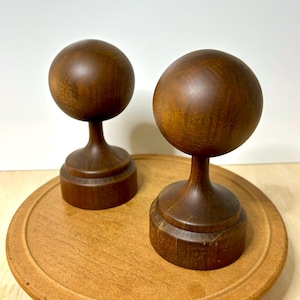 May include: Two wooden decorative objects, each featuring a sphere atop a pedestal base. The objects are a rich, warm brown color, with visible wood grain. They are displayed on a round, light brown wooden surface.