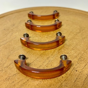 May include: Four amber-colored, curved cabinet pulls with silver screws. The pulls are made of a translucent material, and are arranged in a row on a wooden surface. The pulls have a classic, arched design.