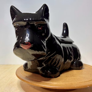 May include: A glossy black ceramic figurine shaped like a Scottish Terrier dog. The dog is depicted in a seated position with pointed ears, a short upright tail, and a smooth, reflective surface. The piece features a removable lid on the back.