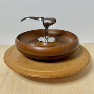 May include: A wooden nut bowl with a built-in chrome nutcracker. The bowl is a rich brown color and sits on a lighter-colored wooden base. The nutcracker is silver and has a curved handle.