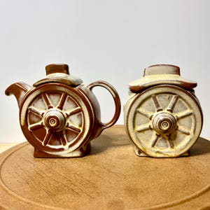 May include: A ceramic teapot and a matching lidded sugar bowl, both featuring a prominent wagon wheel design on their sides. The pieces are finished in a two-tone glaze of deep brown and creamy off-white, with a textured, rustic appearance.