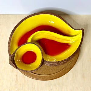 May include: A ceramic divided serving dish with a bright yellow interior and a deep red center. The exterior features a textured brown finish resembling tree bark. The dish has a curved, leaf-like shape with a separate circular compartment.