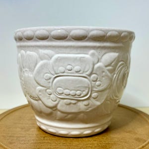May include: A white ceramic flower pot with embossed floral designs. The pot has a rounded shape with a decorative rim and a detailed pattern of flowers and leaves. The pot is sitting on a wooden surface.