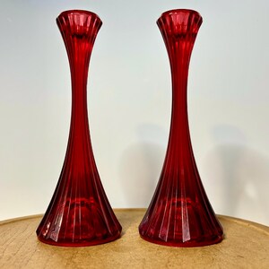 May include: A pair of tall, vibrant red glass candle holders featuring a fluted, vertical ribbed design. Each holder has a wide, flared base that tapers into a slender neck, topped with a circular opening for a candle, crafted from translucent red glass.