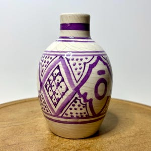 May include: A small, bulbous ceramic vase with a cream-colored base and a visible crackle glaze finish. The surface is decorated with hand-painted purple geometric patterns, including diamonds, dots, and circular motifs around the body.