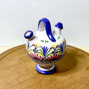 May include: A white ceramic pitcher with a blue handle and spout, adorned with colorful floral patterns in red, yellow, and blue. The pitcher has a small base and a round body, perfect for decorative use.