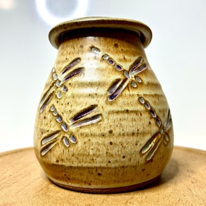 May include: A handcrafted, ceramic jar with a lid. The jar is tan with a speckled finish and features embossed dragonfly designs with purple accents. The jar is sitting on a wooden surface. The jar is a decorative item.