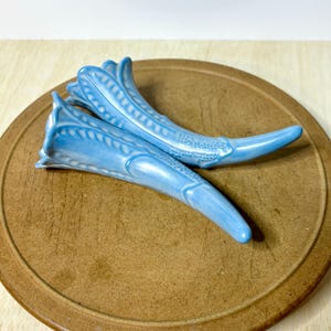 May include: Two light blue ceramic cornucopia-shaped vases with a glossy finish. Each piece features a flared, petal-like opening and a textured, embossed pattern along the curved body, tapering to a smooth, pointed end at the base.