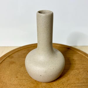 May include: A beige ceramic vase with a rounded base and a long, slender neck. The vase has a speckled texture and sits on a wooden tray. The vase is empty and ready to be filled with flowers or used as a decorative piece.