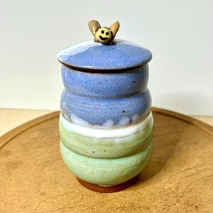 May include: A cylindrical ceramic jar with a lid featuring a small sculpted bee handle. The jar has a smooth glaze transition from light blue at the top to a white band in the middle and a pale green base, with a dark brown exposed clay bottom.