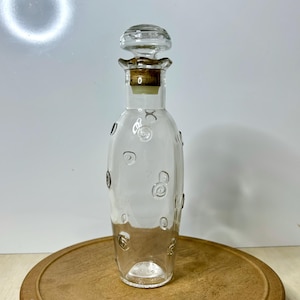 May include: A clear glass bottle with a decorative stopper. The bottle has a cork and a clear glass stopper. The bottle is decorated with raised spiral designs. The bottle is sitting on a wooden surface.