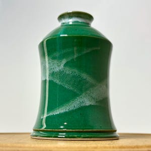 May include: A handcrafted, green ceramic jar with a lid. The jar has a glossy finish with white, abstract markings. The rim and base are a darker green, and the lid has a white accent. The jar is a decorative piece.