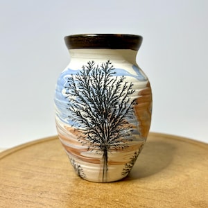 May include: A ceramic vase with a black tree design. The vase has a brown rim and a cream-colored body with blue, brown, and white swirls. The tree is centered on the vase, with detailed branches. The vase is approximately 6 inches tall.