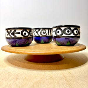 May include: Three small ceramic bowls with a rounded shape. Each bowl features a white upper band decorated with black hand-painted abstract symbols and circular motifs, while the lower section is a deep blue with green triangular accents.