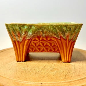 May include: A rectangular ceramic planter featuring a vibrant orange base with a geometric star pattern and a contrasting light green drip glaze along the top rim. The vessel is supported by four sturdy, integrated feet at the corners.
