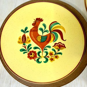 May include: Round, yellow plate with a brown rim. The plate features a colorful rooster design in the center, surrounded by floral accents. The rooster is red, with green, yellow, and brown details. The floral accents are yellow, red, and green.