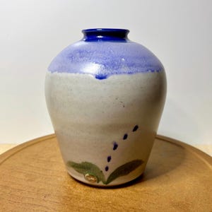 May include: A small ceramic vase with a rounded body and a narrow neck. The upper portion is glazed in a vibrant blue, transitioning into a light, speckled off-white base. A small green leaf design with blue dots is painted near the bottom.