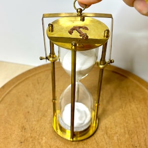 May include: A vintage brass hourglass with a nautical anchor emblem. The hourglass features a clear glass chamber filled with white sand, encased in a brass frame. The design includes a circular top and bottom, connected by vertical brass supports. The hourglass is a decorative item.