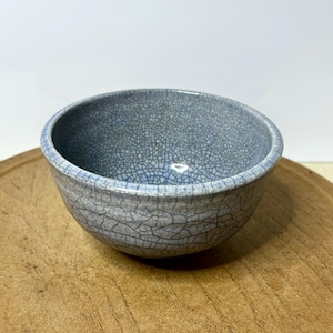 May include: A small, round, ceramic bowl with a light blue, crackled glaze. The bowl sits on a wooden surface. The interior and exterior of the bowl have a textured appearance, with a network of fine lines.