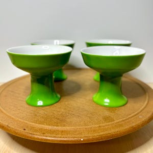May include: Four green ceramic dessert cups with white interiors are arranged on a wooden serving tray. The cups have a pedestal base and a wide, shallow bowl. The bright green color contrasts with the white interior and the natural wood tone of the tray.